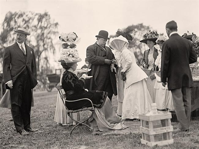 Nellie Taft, seated, having her cake and eating it too. Will stands engaging with lawn party guests on a chilly day, a big glass water container informally plopped near them. Their daughter Helen stands, in large black hat, third from the right.